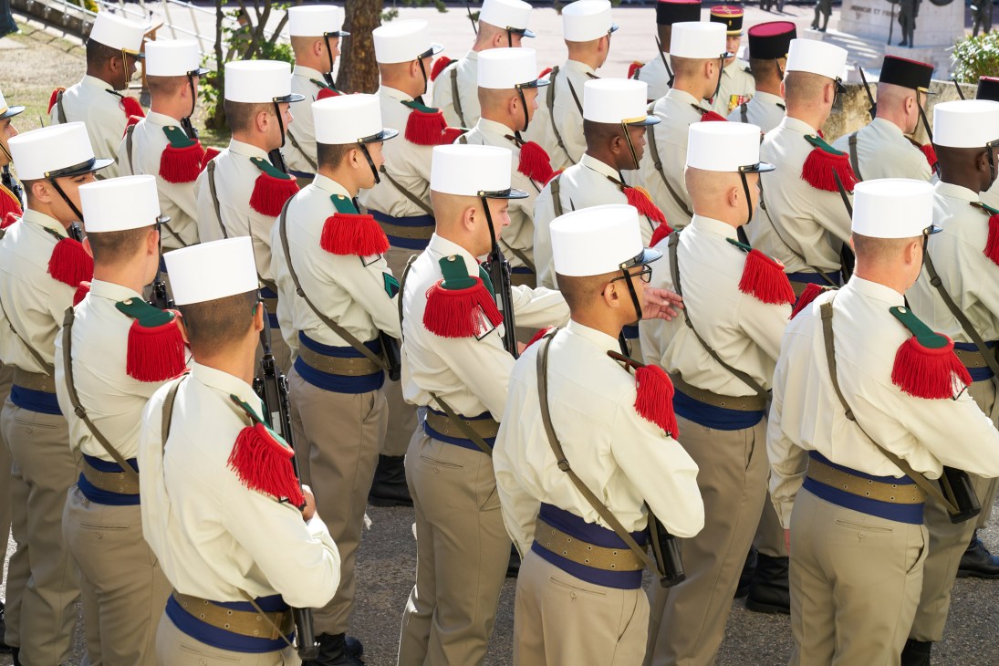 French Foreign Legion soldiers in formal dress uniform featuring white képi blanc caps, cream uniforms, blue sashes, and red épaulettes.