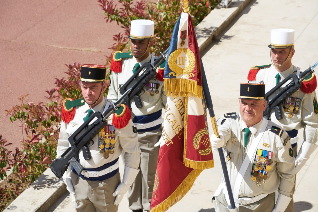 French Foreign Legion soldiers in dress uniform carrying a regimental flag with gold embroidery and red tassel.