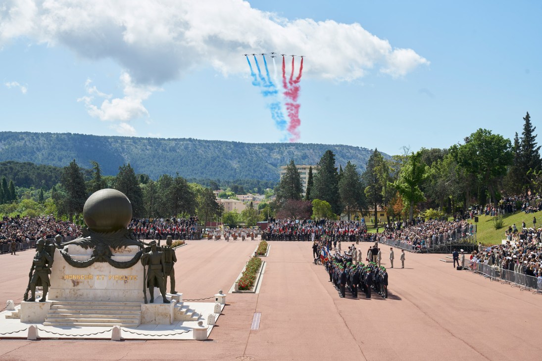 French military jets perform a flyover trailing blue, white and red smoke above a large parade ground during Camerone Day ceremony.