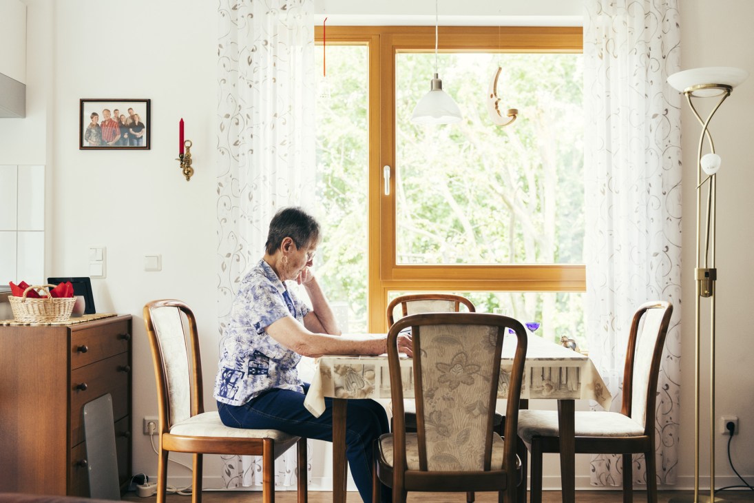 Woman sitting at dining table in bright living room with large windows overlooking greenery.
