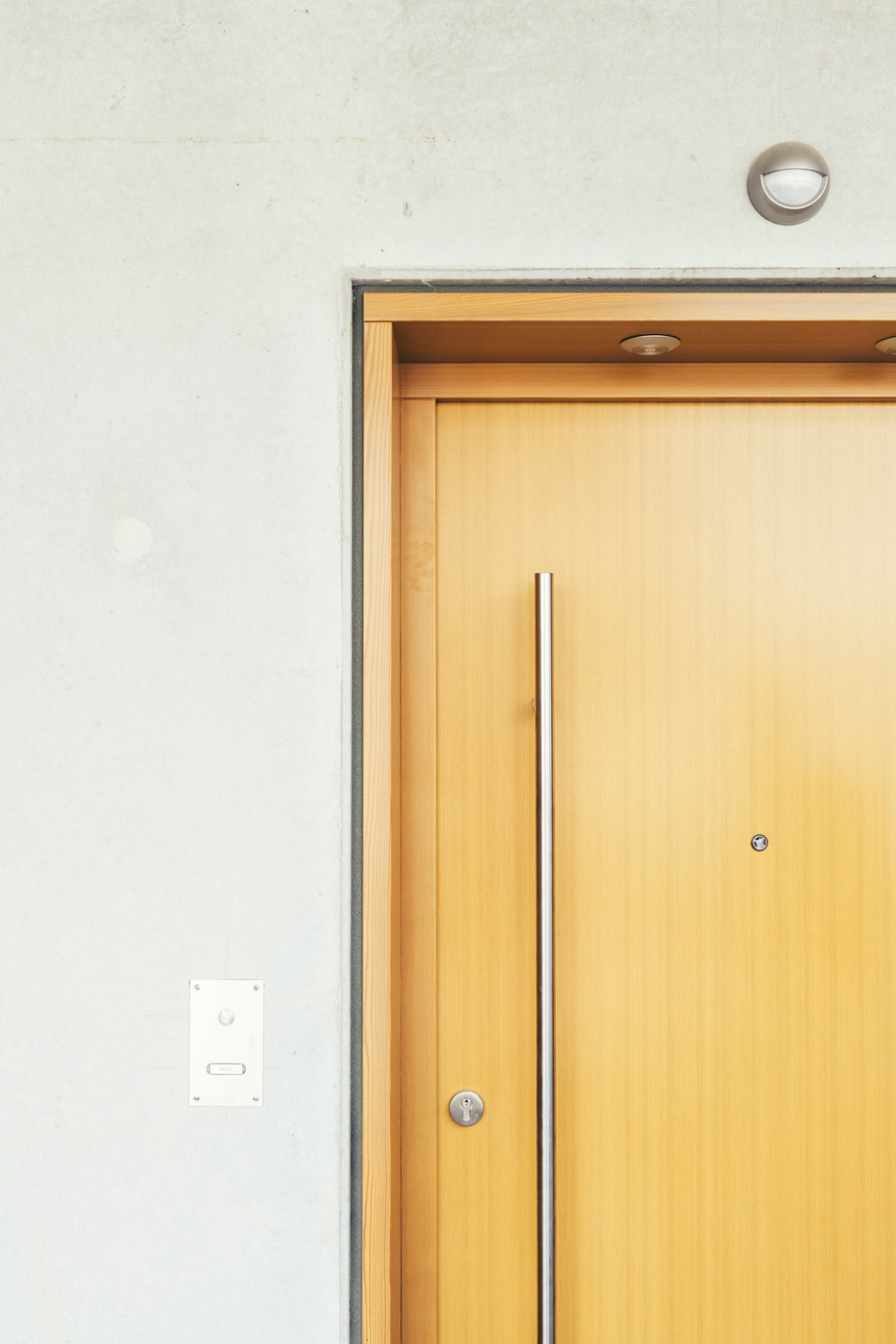 Light wood apartment door with metal handle and security lock against white wall with circular light fixture above.
