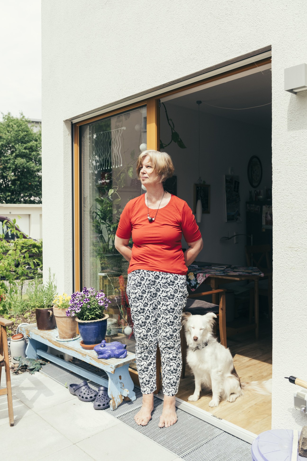 Woman in red shirt and patterned trousers standing in doorway with white dog beside her.