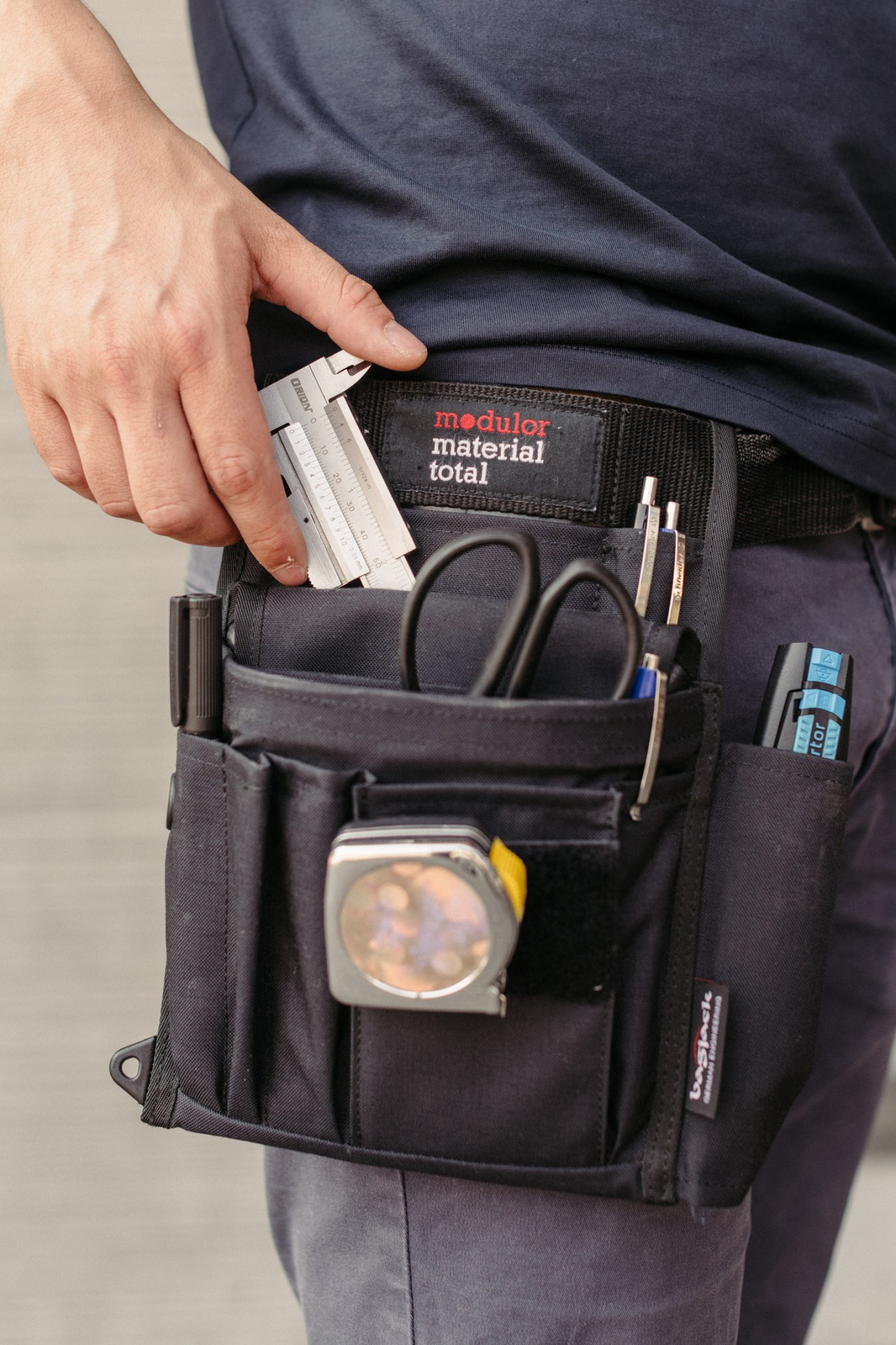 A person wearing a dark apron with Modulor branding holds a ruler, displaying the apron's contents including scissors, pens, and tape.