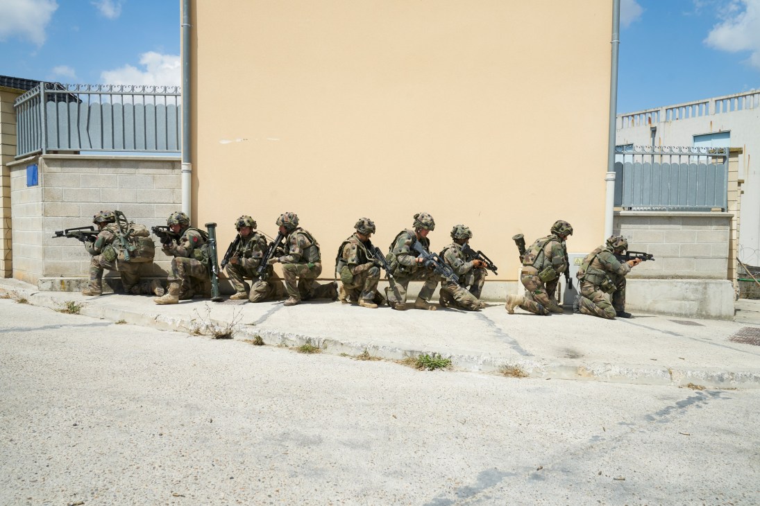 Armed legionnaires in camouflage uniforms defend positions along a concrete wall during urban warfare training.