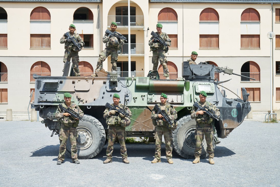 Legionnaires in combat gear wearing green woollen berets standing on and around a military truck in a town square.