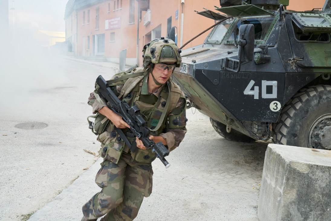 Legionnaire in camouflage uniform and helmet carrying rifle during urban warfare training exercise with military vehicle in background