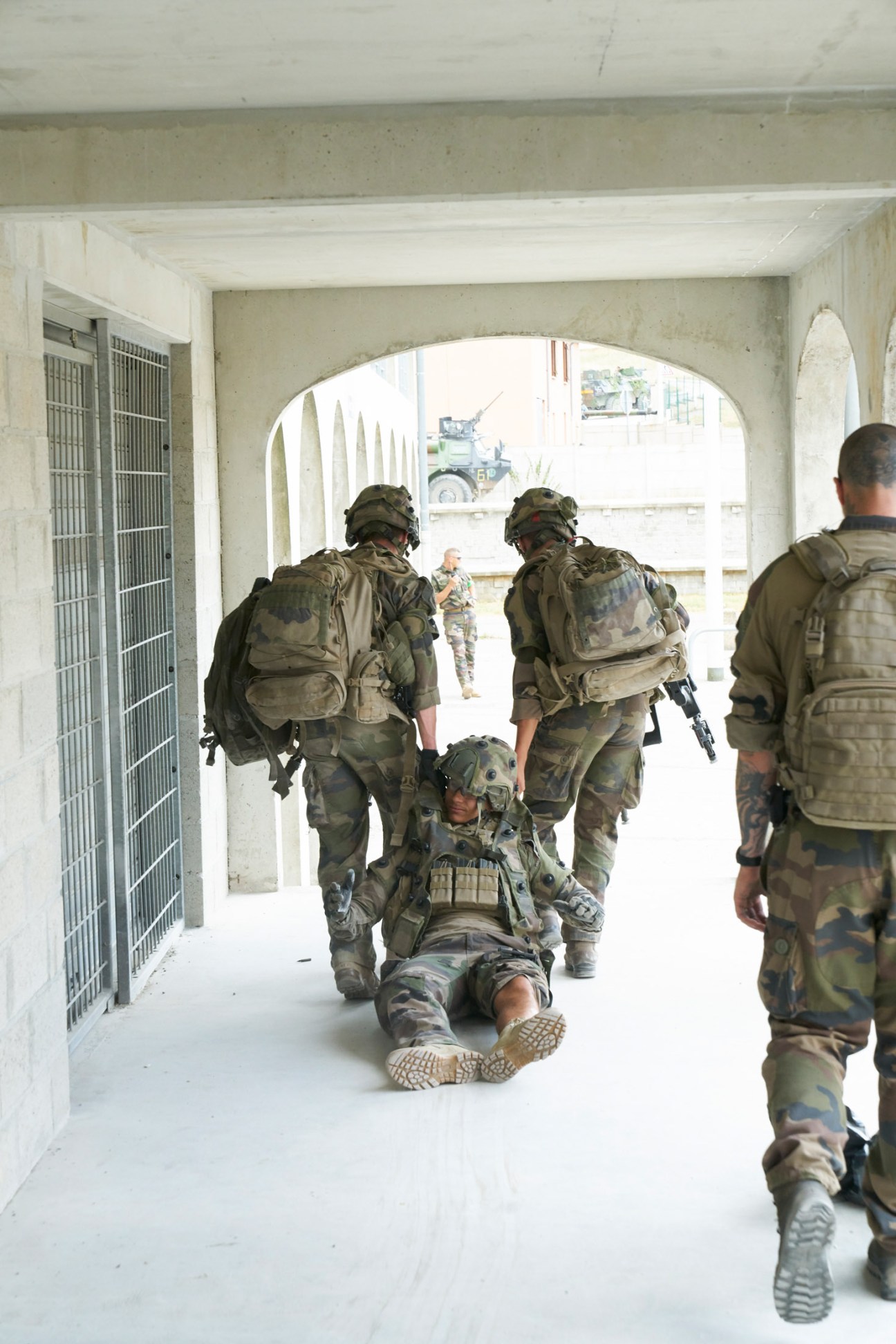 Soldiers in camouflage uniforms dragging an injured comrade to safety during a training exercise in a concrete corridor.