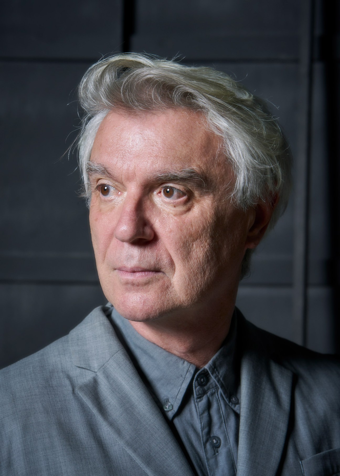 David Byrne, an older man with white hair, wearing a grey button-up shirt against a dark background.