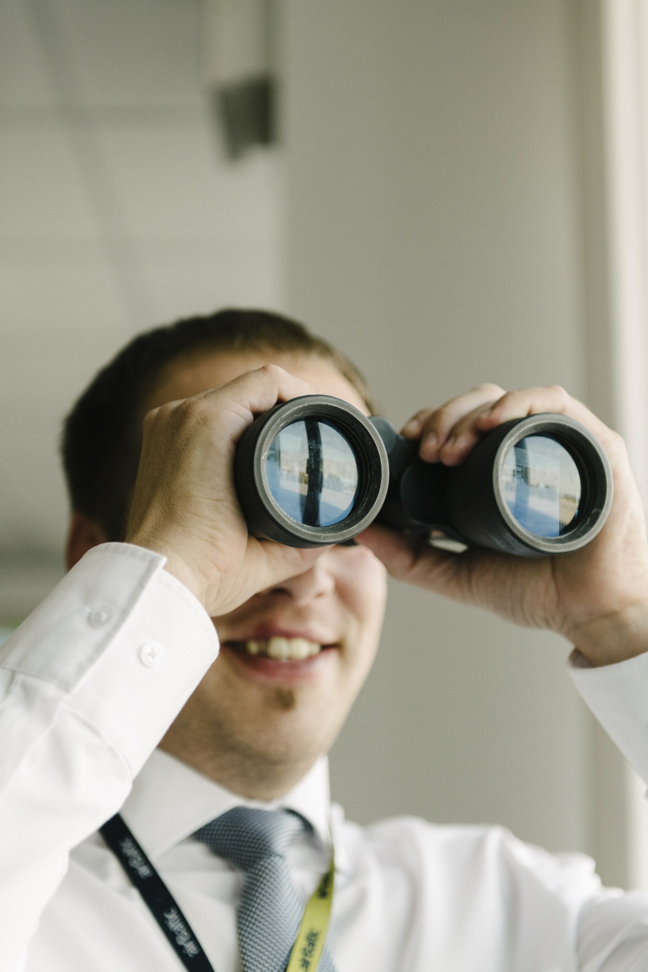 Man in white shirt and tie holding binoculars up to his eyes, smiling indoors