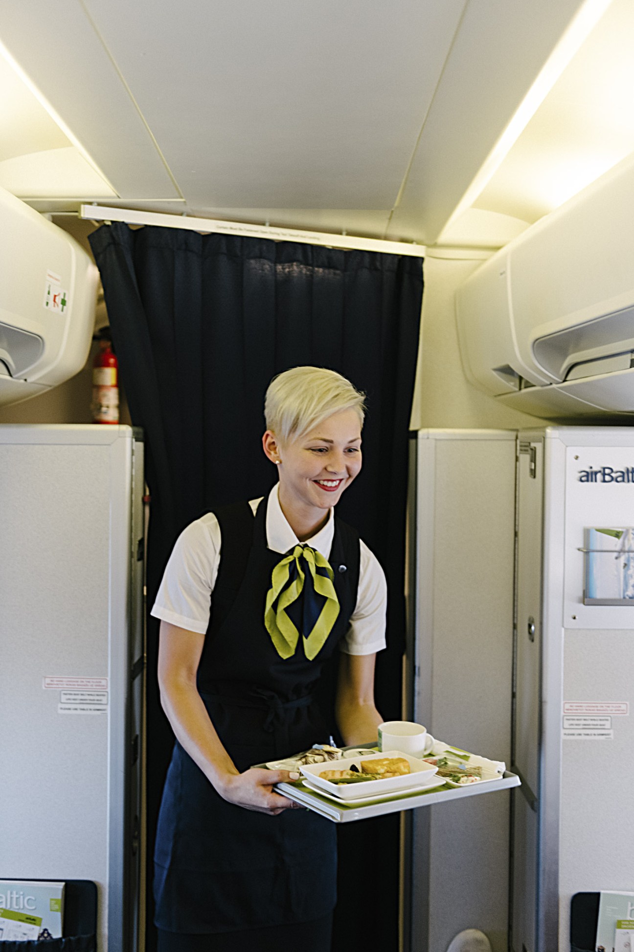 AirBaltic flight attendant serving food in aircraft cabin wearing uniform with yellow scarf