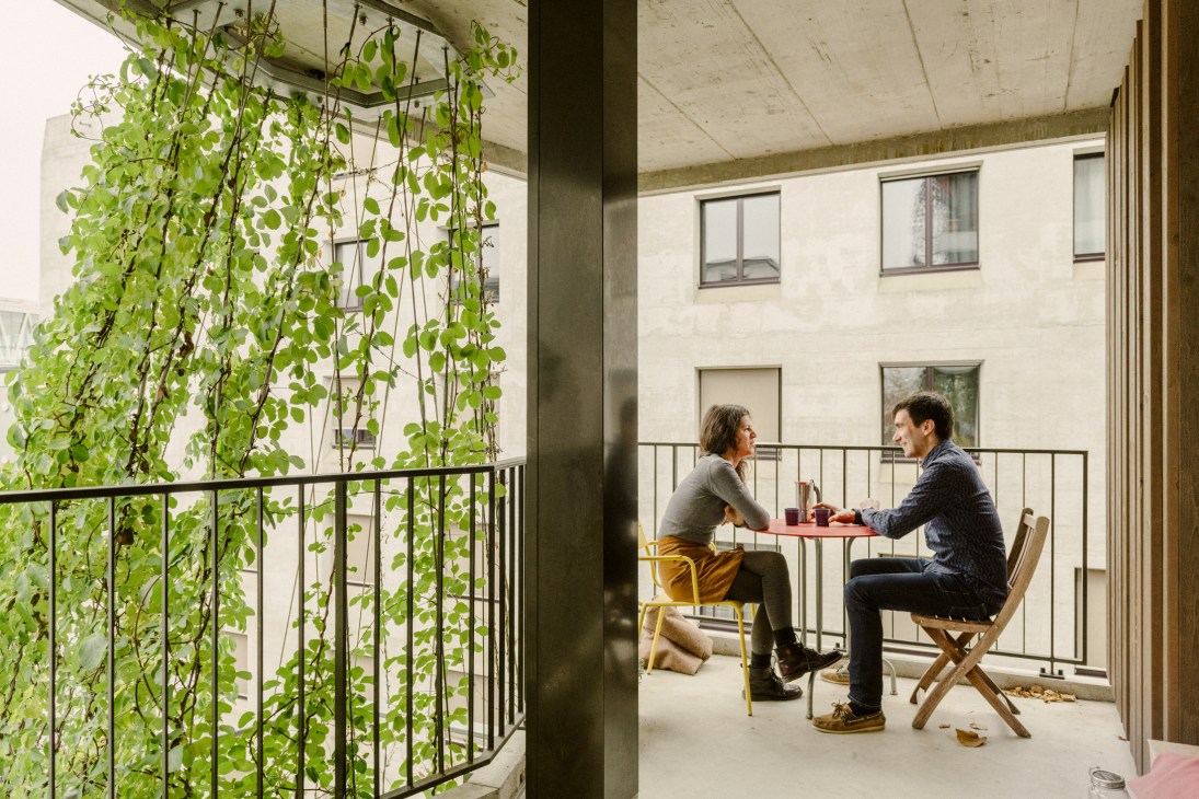 Two men sit facing each other at a small table on a concrete balcony with climbing ivy and residential buildings in the background.