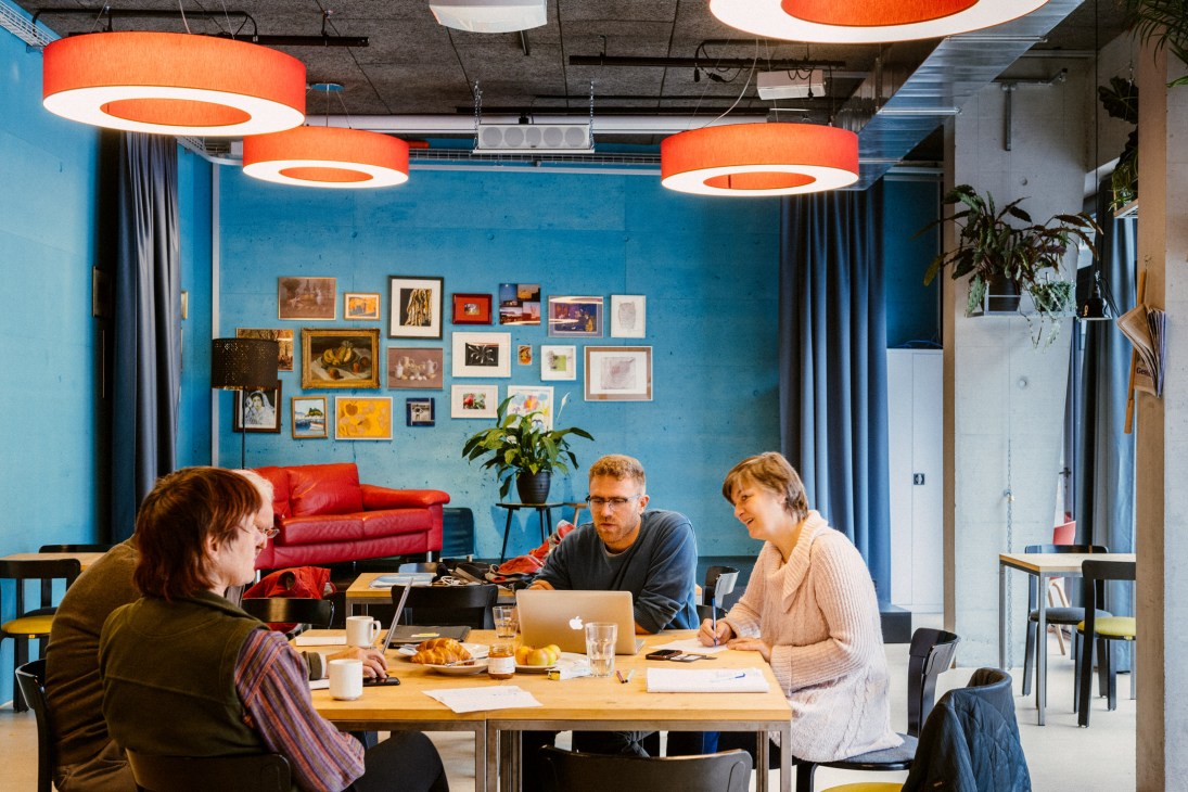 Three residents discuss around a wooden table in a bright blue meeting space with orange pendant lights.