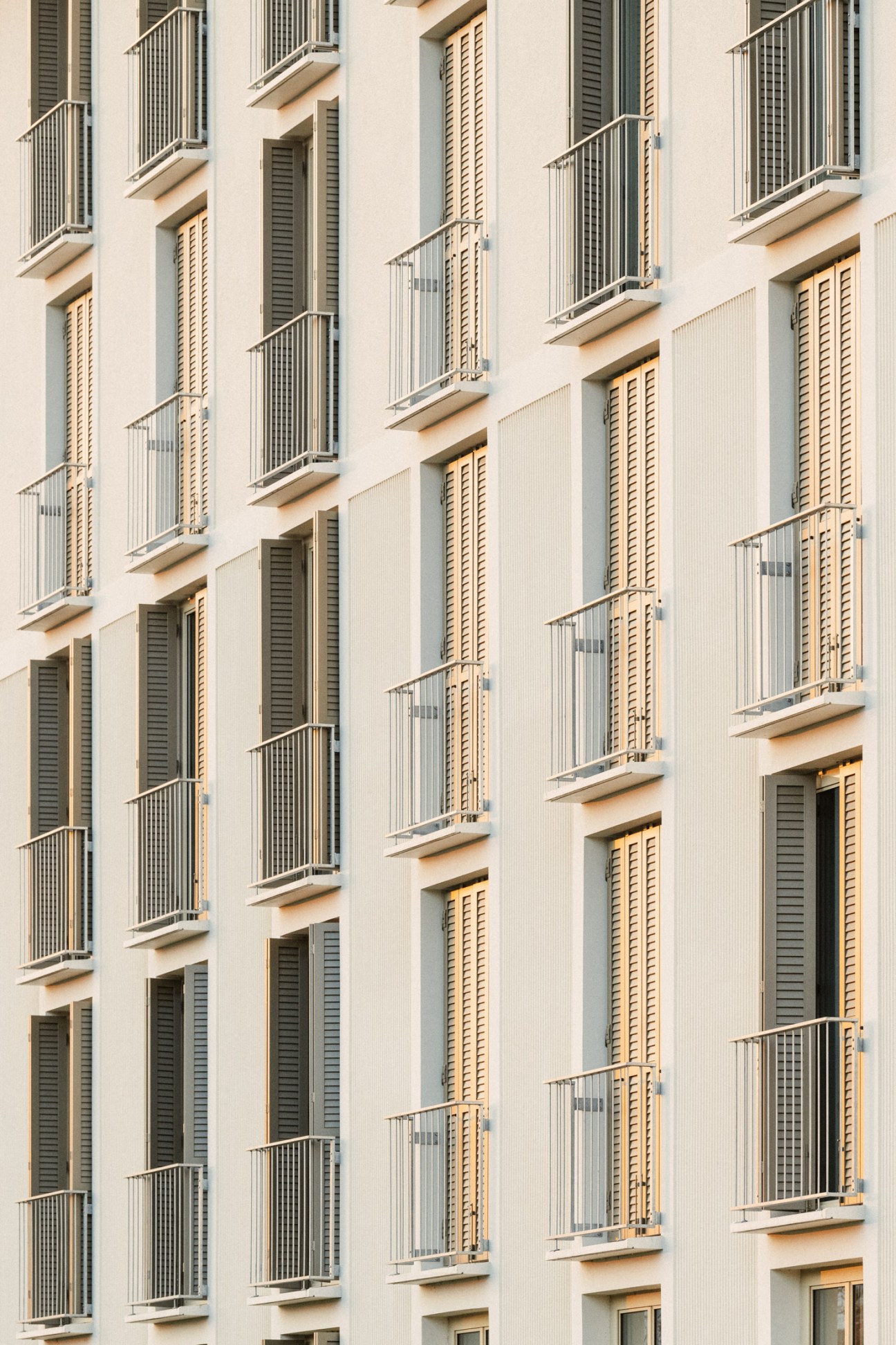 Cream-colored residential building facade with rows of windows, wooden shutters and metal railings on small balconies.
