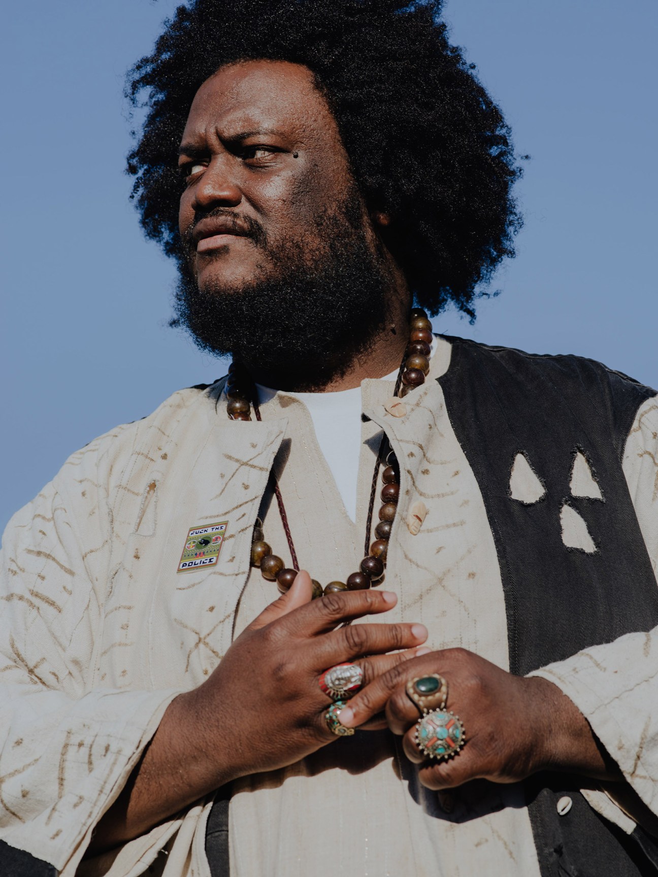 Kamasi Washington wears an African-inspired cream jacket with beaded necklace against a blue sky background.