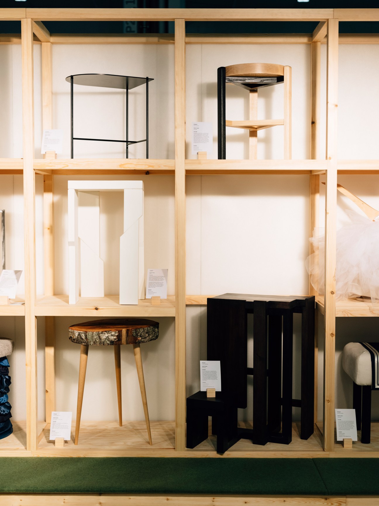 A wooden display unit showcasing various furniture pieces including stools, shelving units, and black sculptural objects with product information cards.