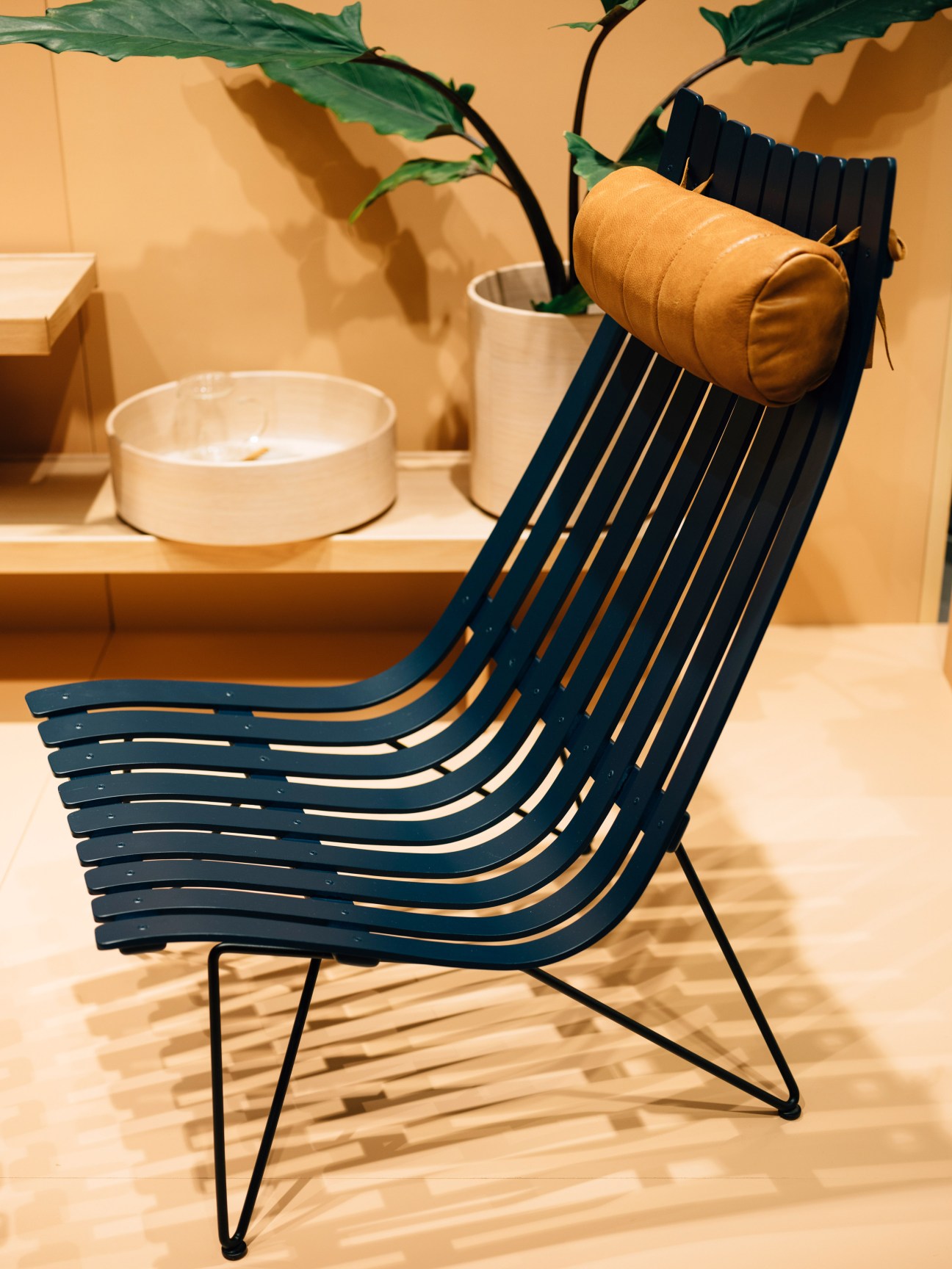 Navy blue slatted chair with tan leather headrest pillow on black metal frame, displayed in minimalist setting with potted plant and ceramic vessels.