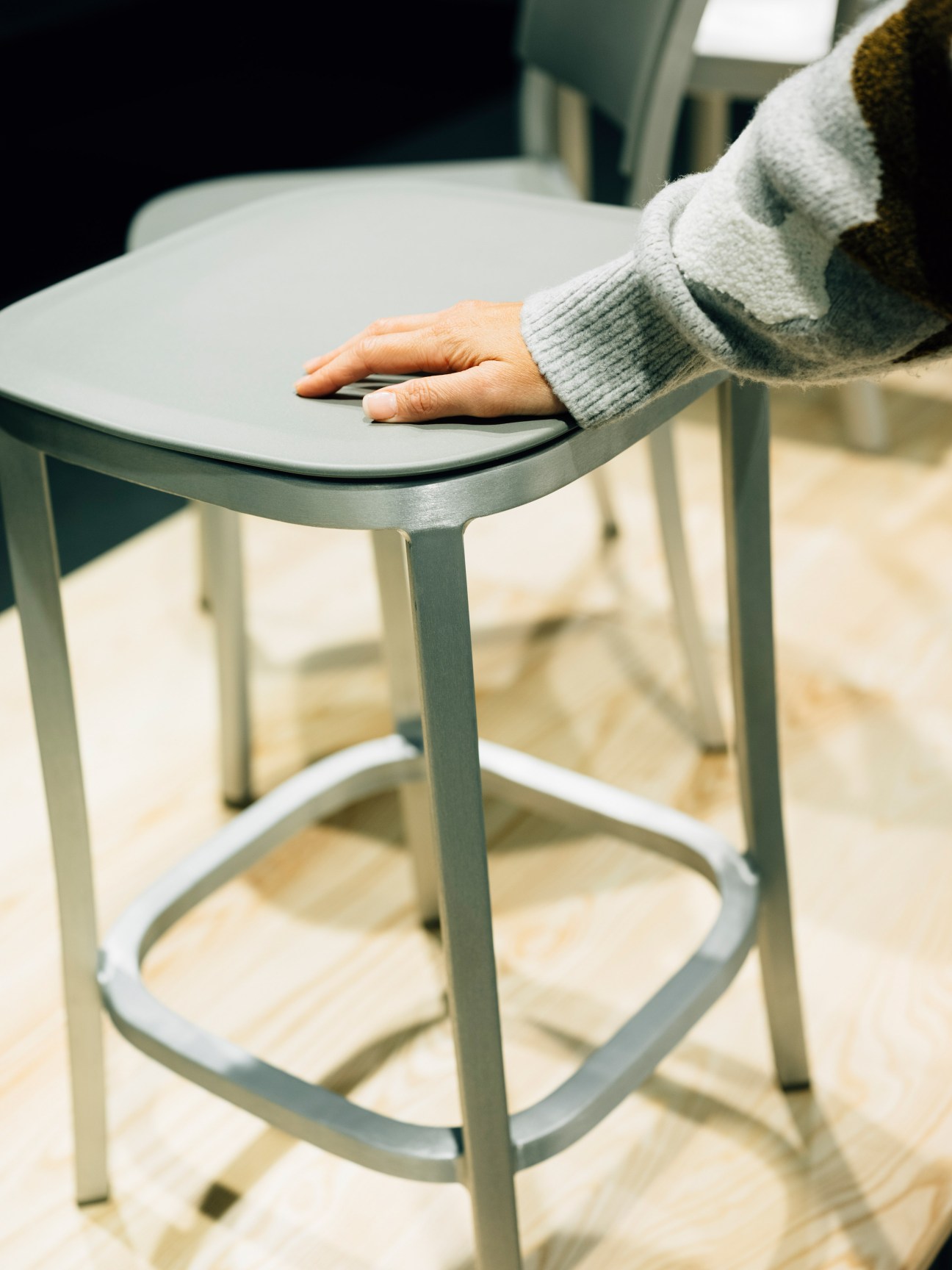 A person's hand touches the seat of a pale green Emeco stool with curved edges and metal legs.