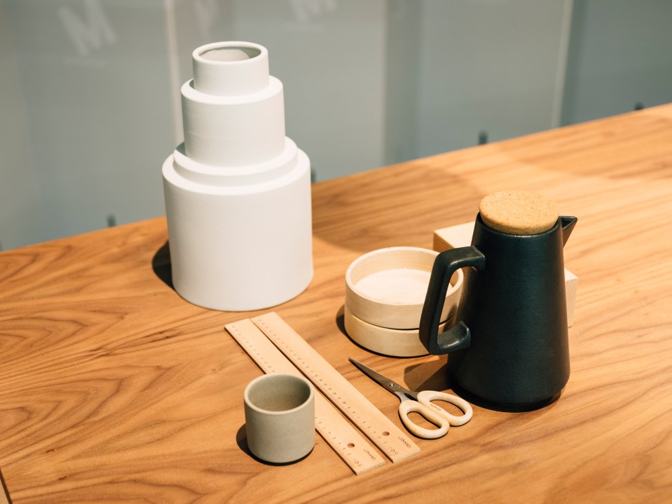 Minimalist ceramic and wooden homeware items arranged on a wooden table, including stacked white vessels, a black pitcher, small cups, wooden utensils, and scissors.