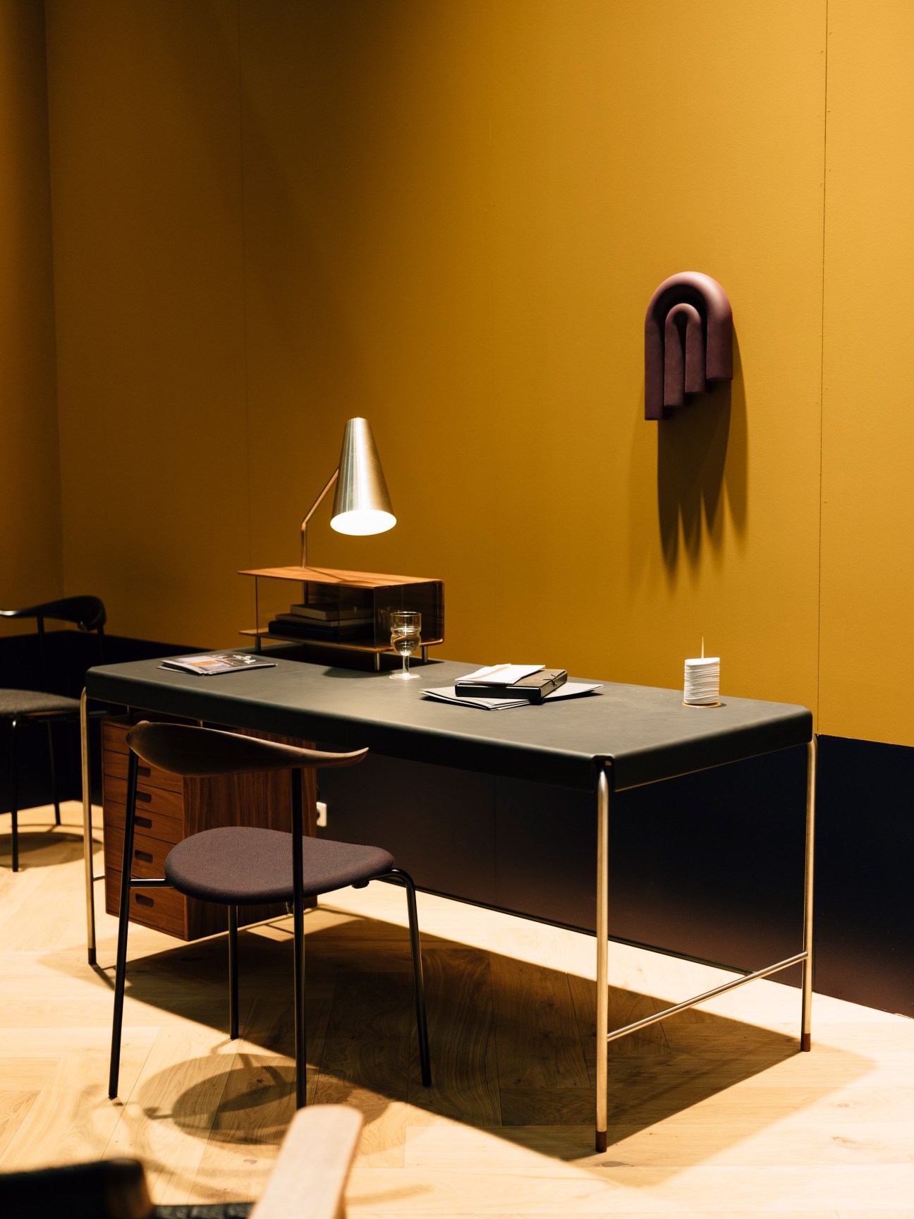 Minimalist desk setup with black table, wooden chair, pendant lamp, and burgundy wall sculpture against mustard yellow wall.