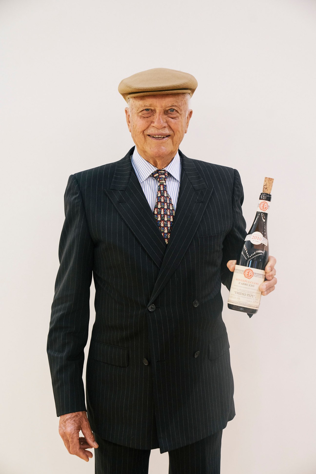 Winemaker Emidio Pepe in a pinstriped blazer holding a wine bottle against a white background.