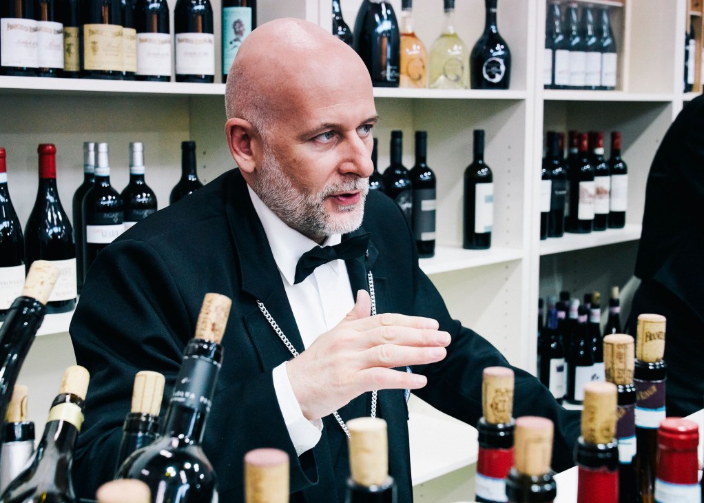 Man in formal attire speaking at a wine exhibition stand with bottles displayed on shelves behind him.
