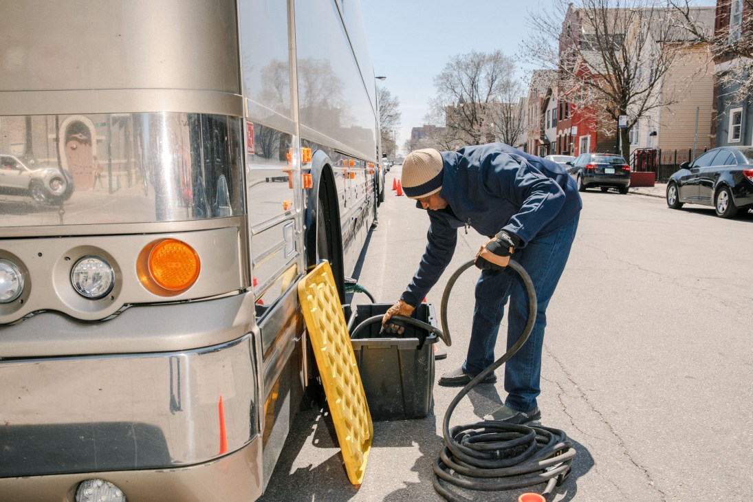 A tour bus driver connects a large black hose to the bus exterior on a city street.