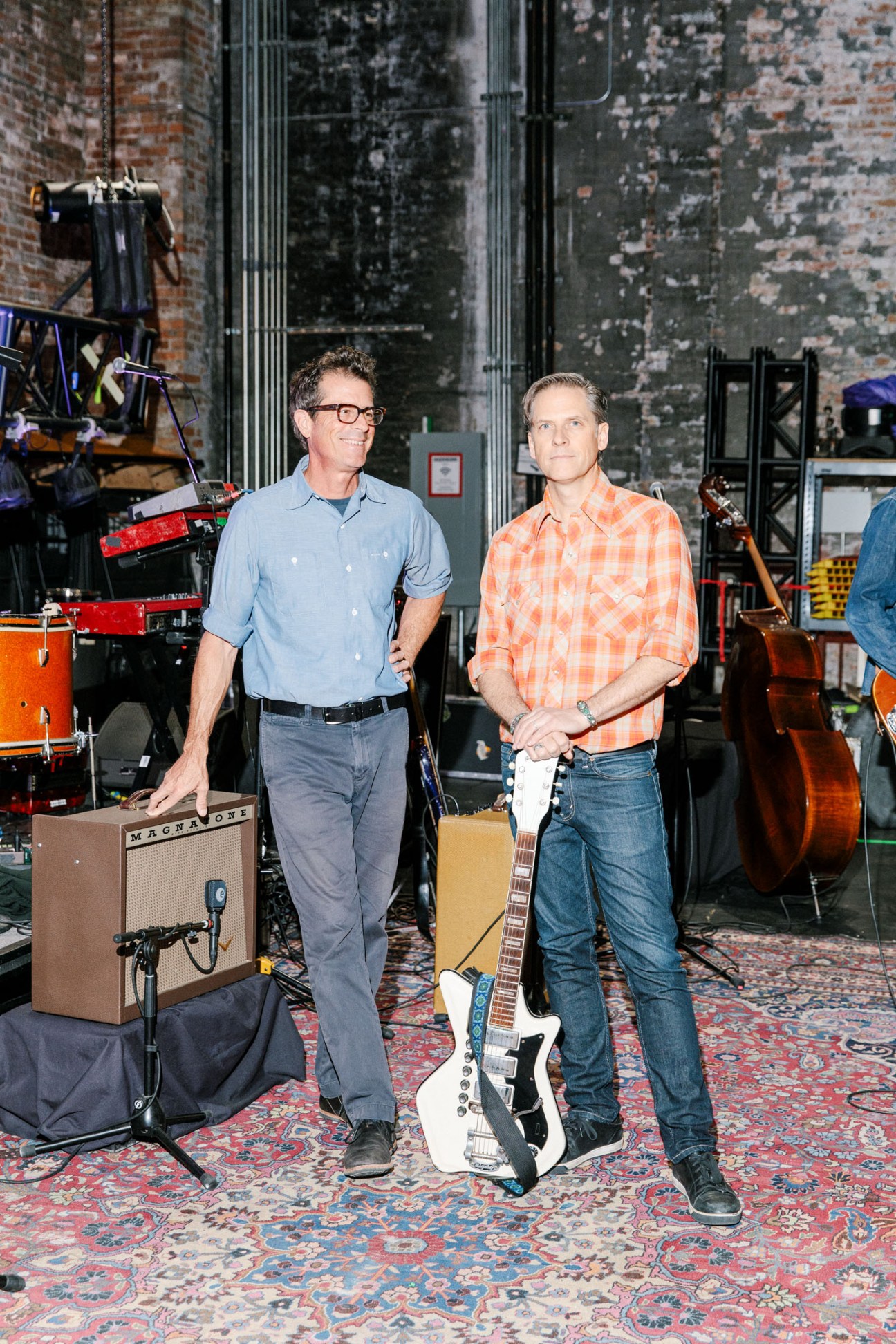 Calexico co-founders John Convertino and Joey Burns stand in a greenroom with musical instruments and equipment behind them.