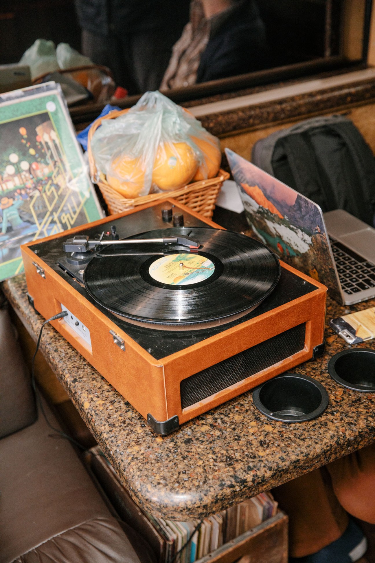Orange vintage record player with vinyl on a granite countertop inside a tour bus, surrounded by magazines and a basket of oranges.