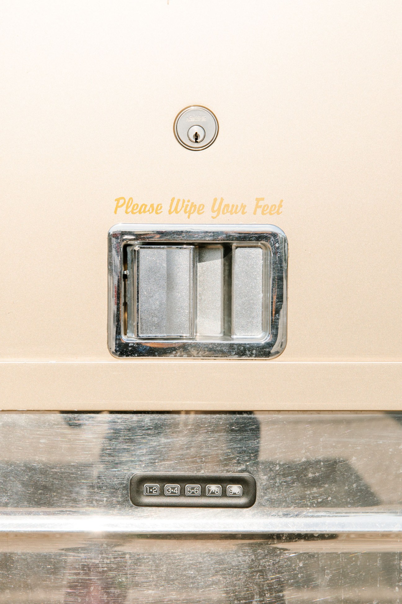 A tour bus entrance with a deadbolt lock above and a mudroom with sliding glass doors below, with yellow text reading 'Please Wipe Your Feet'.