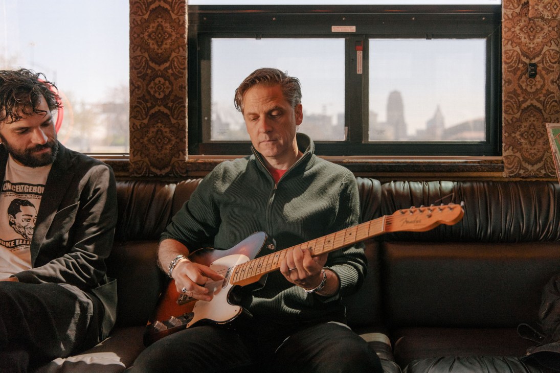 Musician Joey Burns plays guitar on a tour bus with city skyline visible through windows.