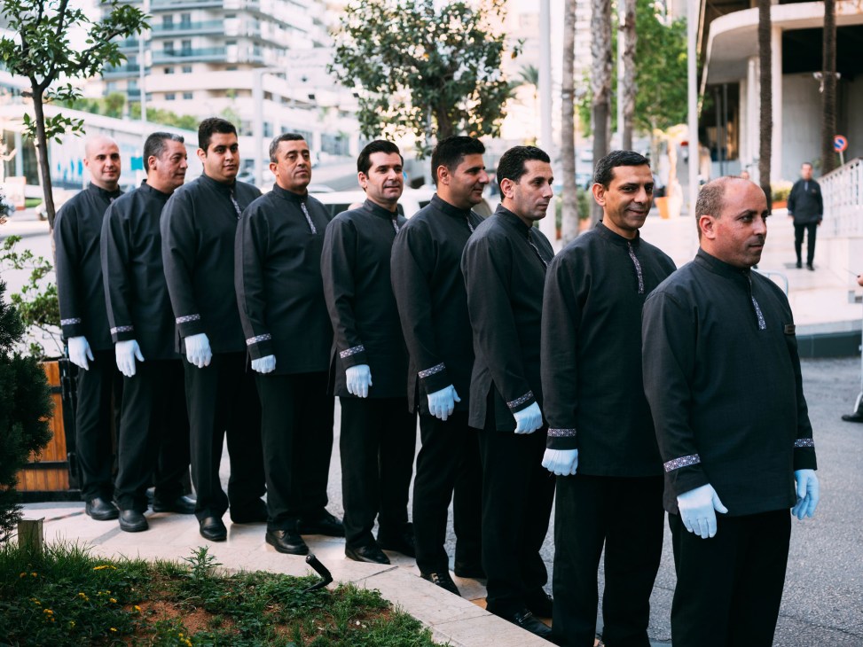 Row of valets in black uniforms with white gloves standing on a Beirut street.