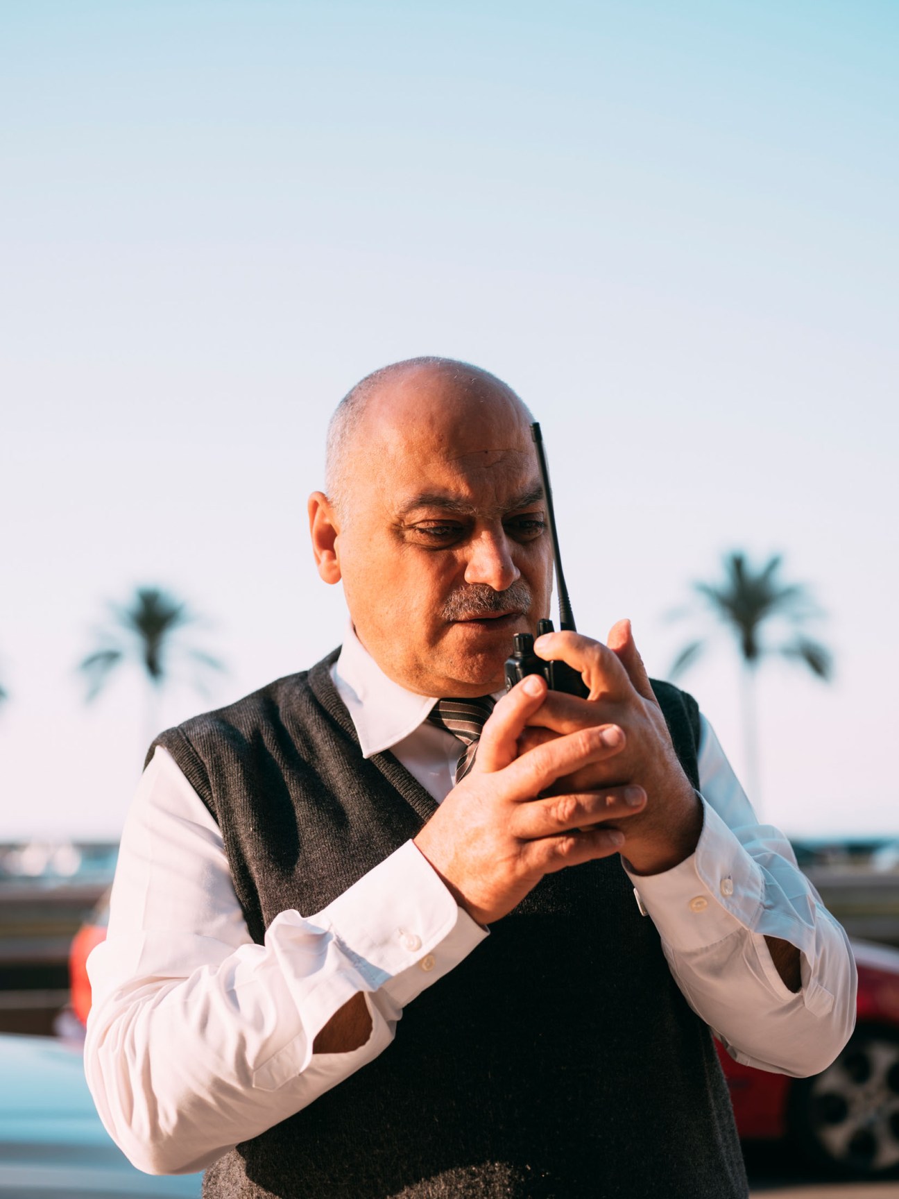 Man in formal attire speaking into a walkie-talkie with palm trees and waterfront in background.