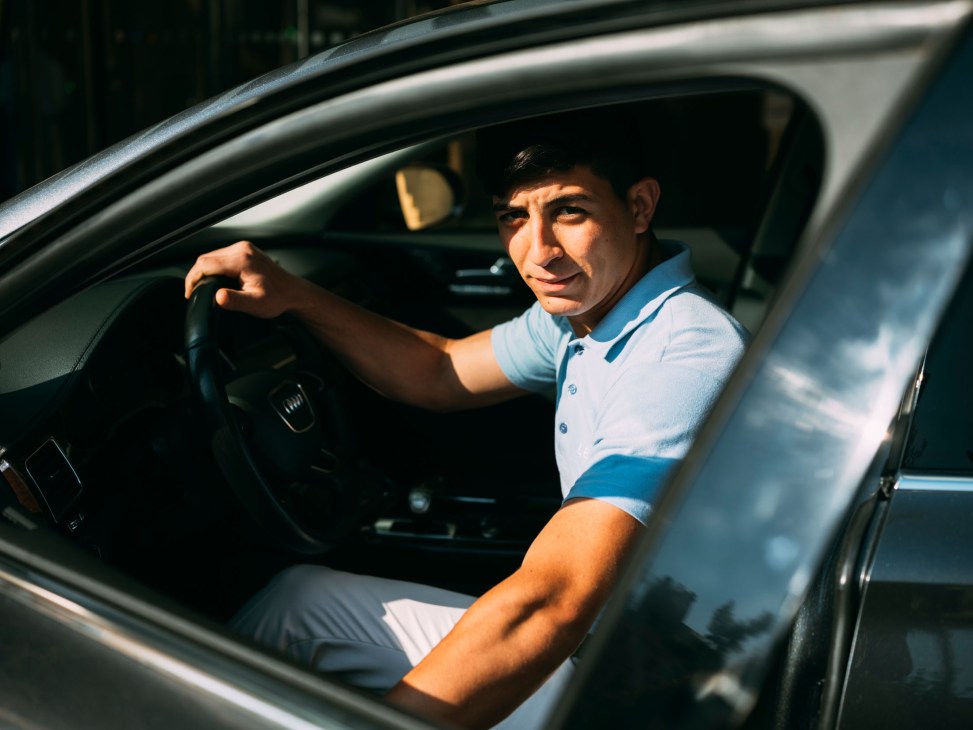 A valet parking attendant in a light blue polo shirt sits behind the steering wheel of a car.
