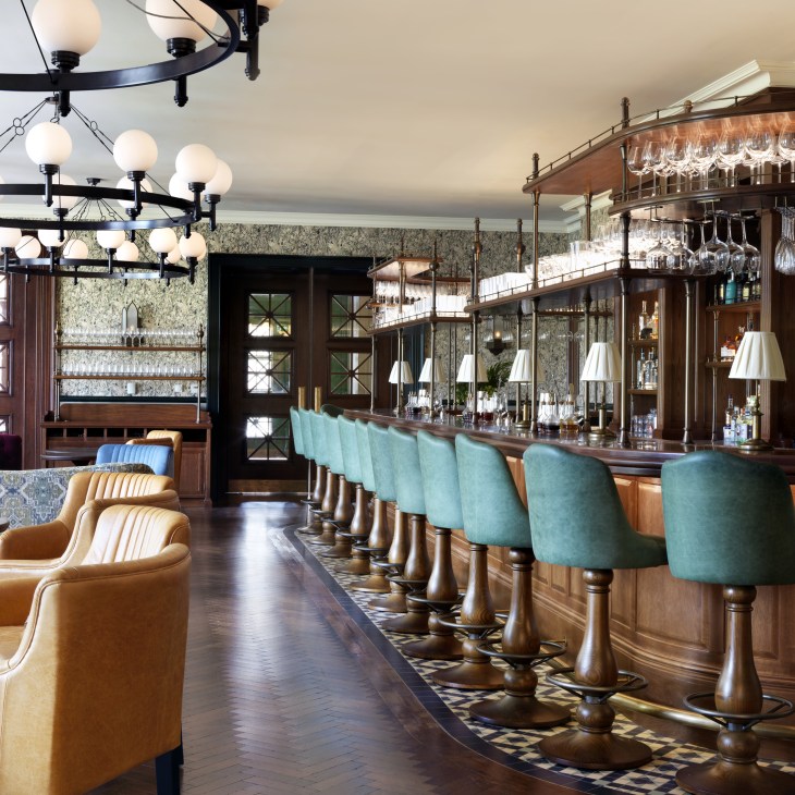 Hotel bar interior with turquoise upholstered bar stools, wooden counter, and contemporary lighting fixtures.
