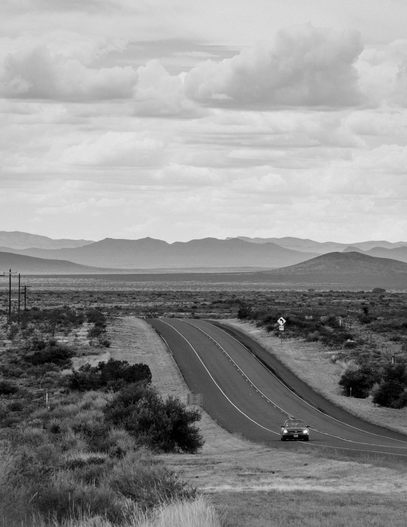 A lone car drives along an empty two-lane road through desert landscape with mountains in the distance