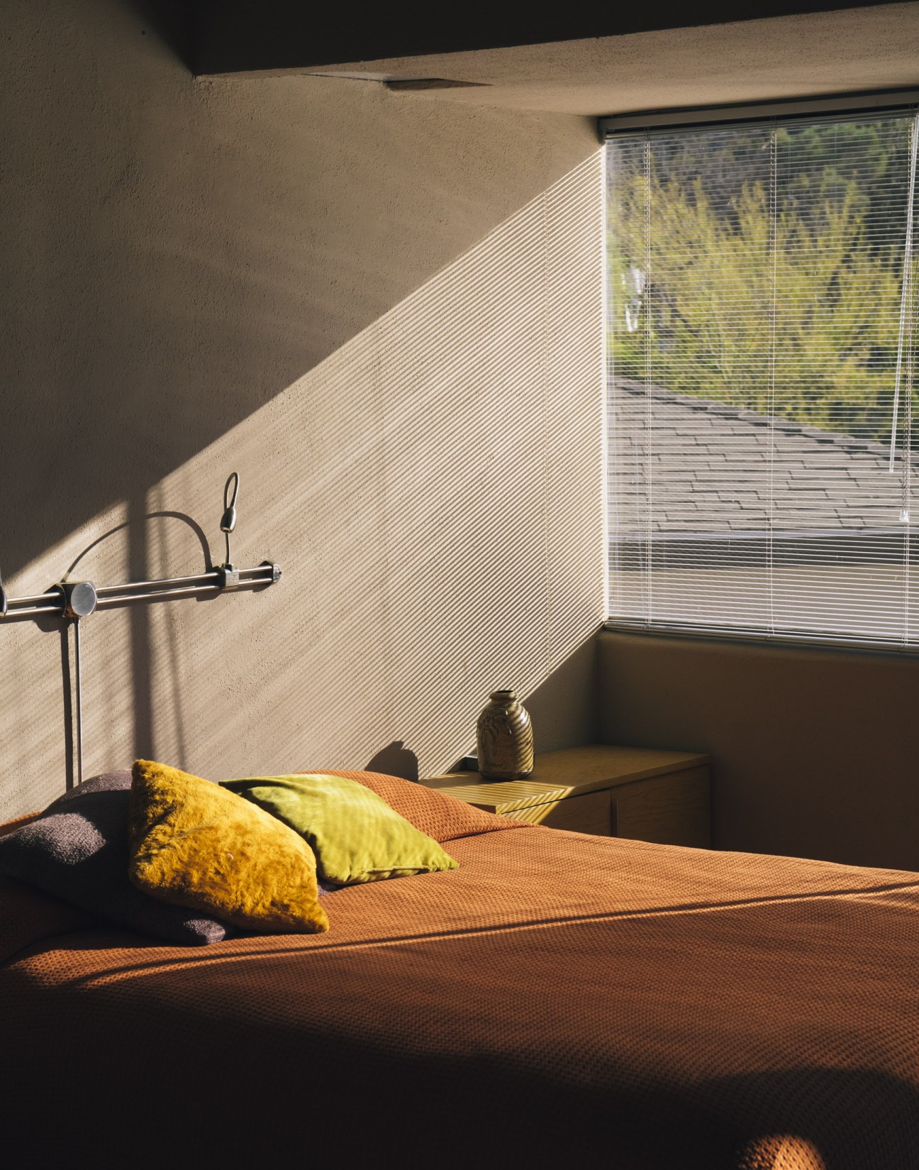 Minimalist bedroom with orange bed, yellow and green pillows, metal bedside lamp, and slatted window blinds overlooking garden.