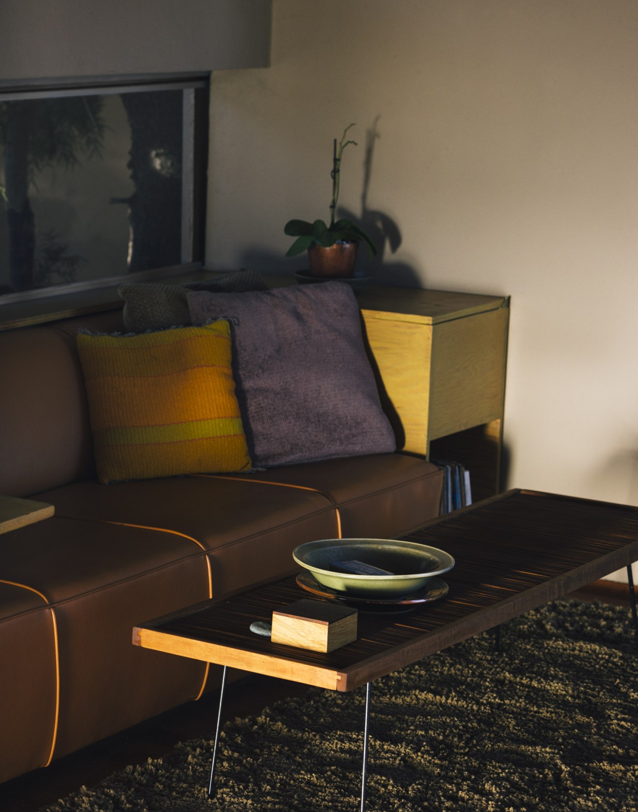 Mid-century modern living room with brown leather sofa, colorful pillows, yellow storage unit, and wooden coffee table near a window.