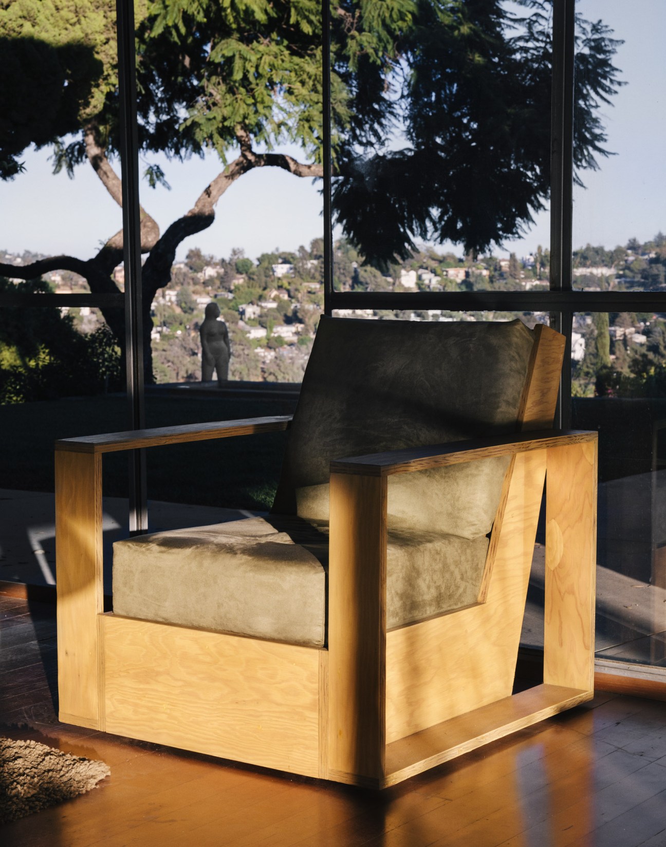 Plywood chair in the style of Schindler with wooden frame and upholstered seat, positioned by large windows overlooking Los Angeles hillside.