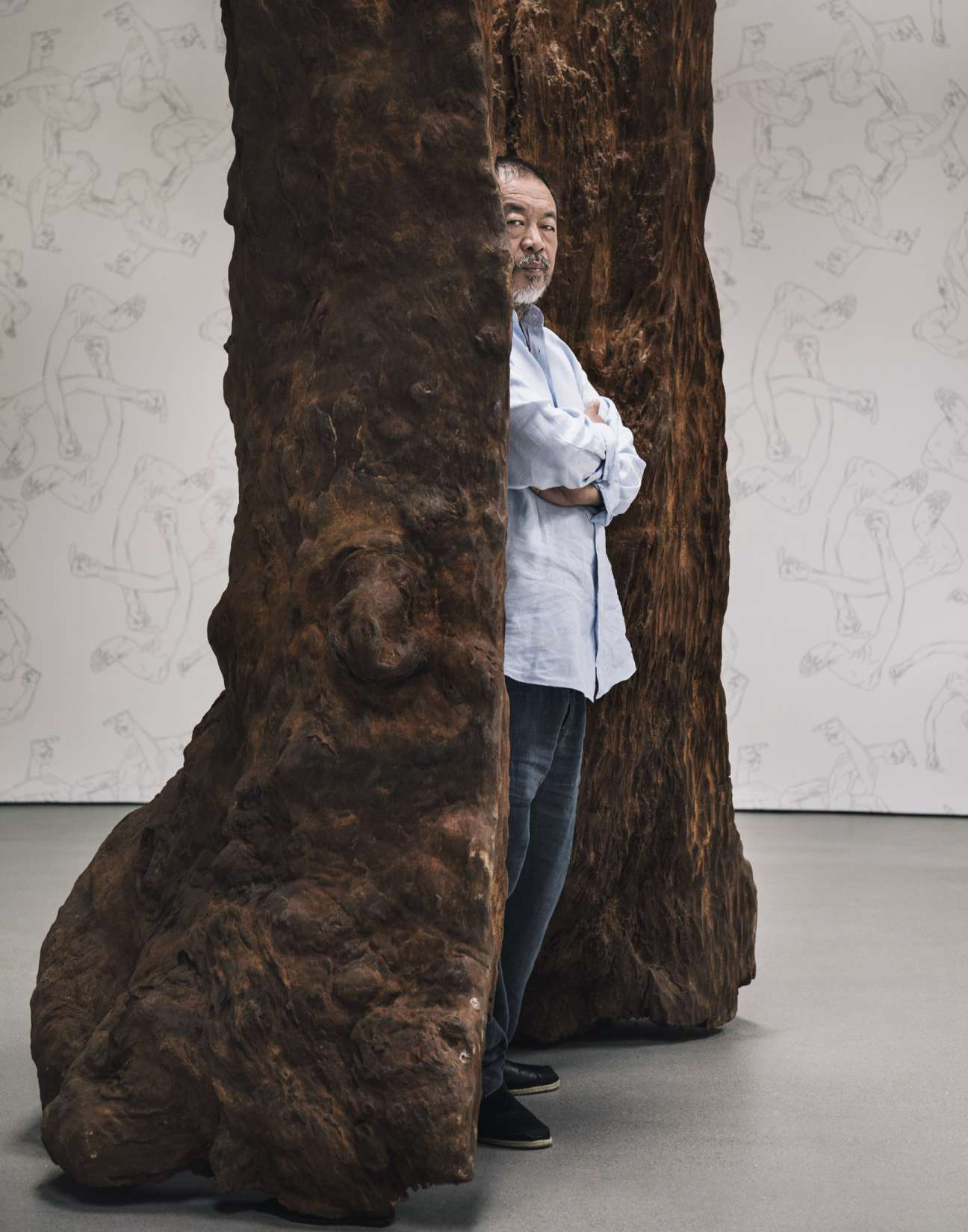 Ai Weiwei standing between two large wooden sculptural forms wearing a light blue shirt