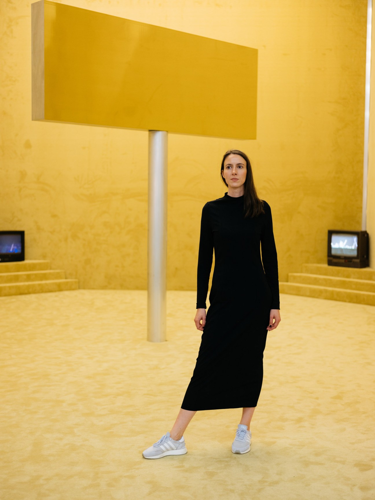 Marie Dessuant stands in a bright yellow installation space with vintage televisions visible on the walls.