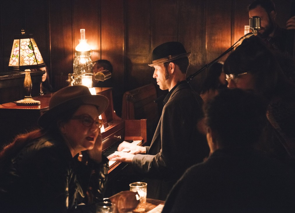 Man in hat playing piano in dim speakeasy with vintage lamps and patrons at The Varnish bar.