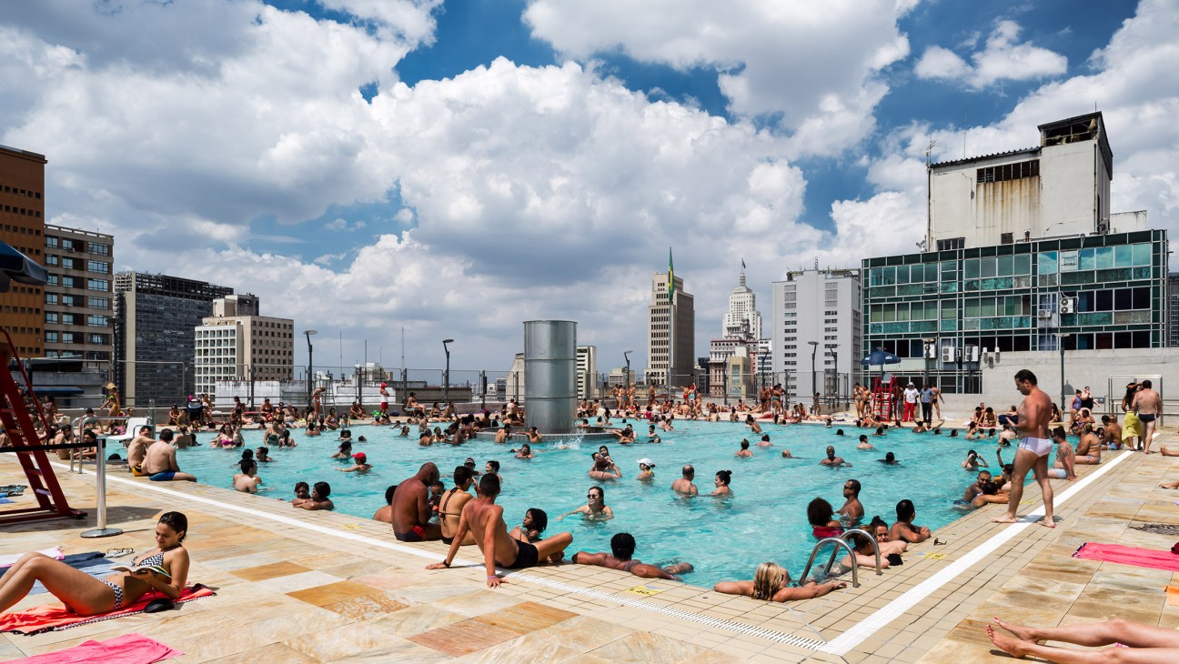 Hundreds of people swim and sunbathe at the rooftop pool of Sesc 24 de Maio in São Paulo with city skyline behind.
