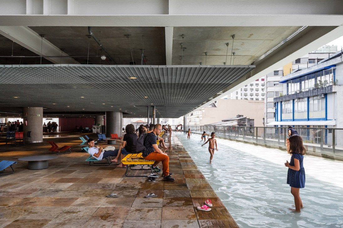 Children and adults enjoy a shallow paddling pool beneath a concrete brutalist structure at Sesc 24 de Maio in São Paulo.