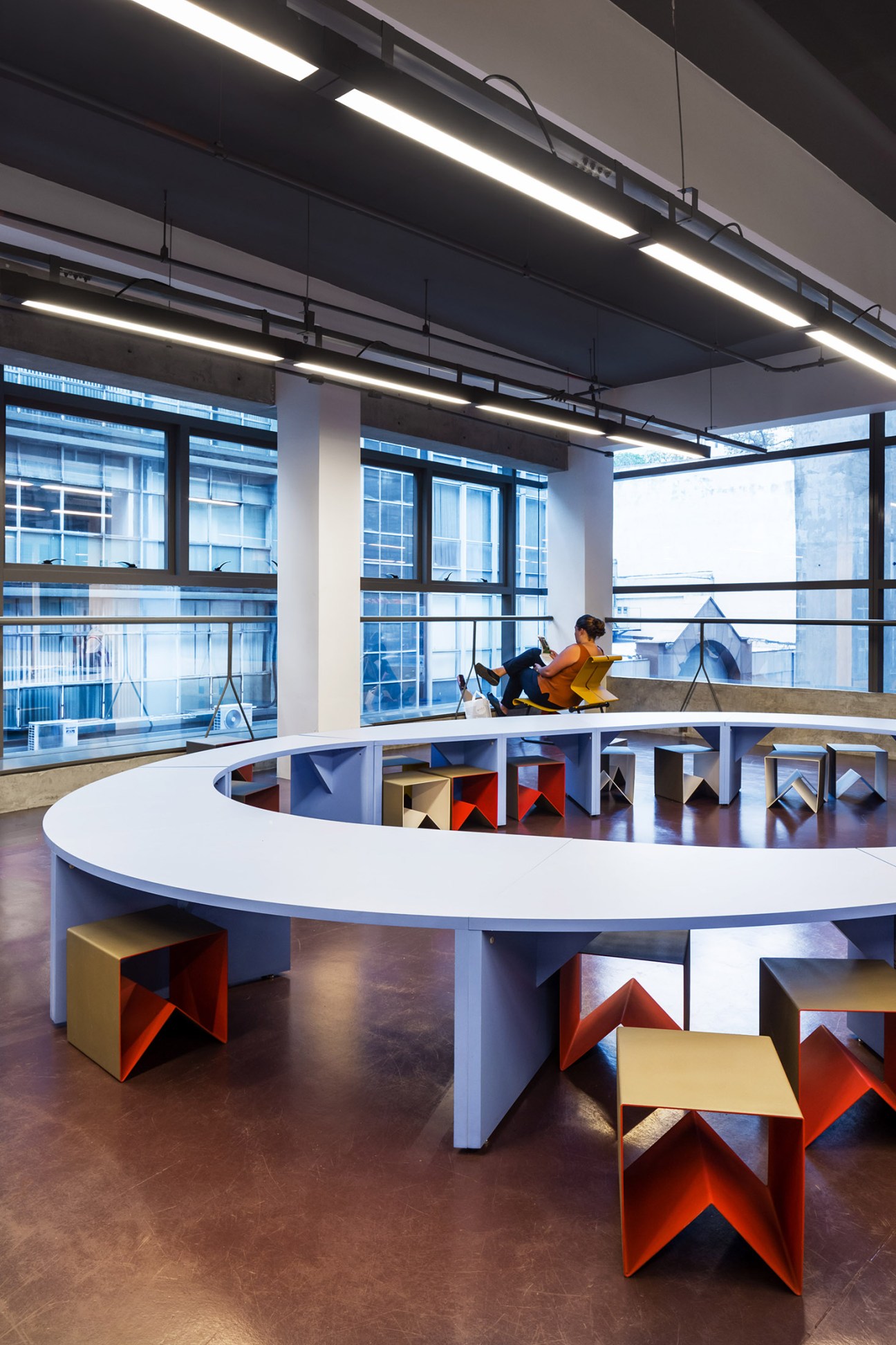 Modern library seating area with colorful angular chairs in red, yellow, and blue arranged around white circular tables with large windows overlooking São Paulo.