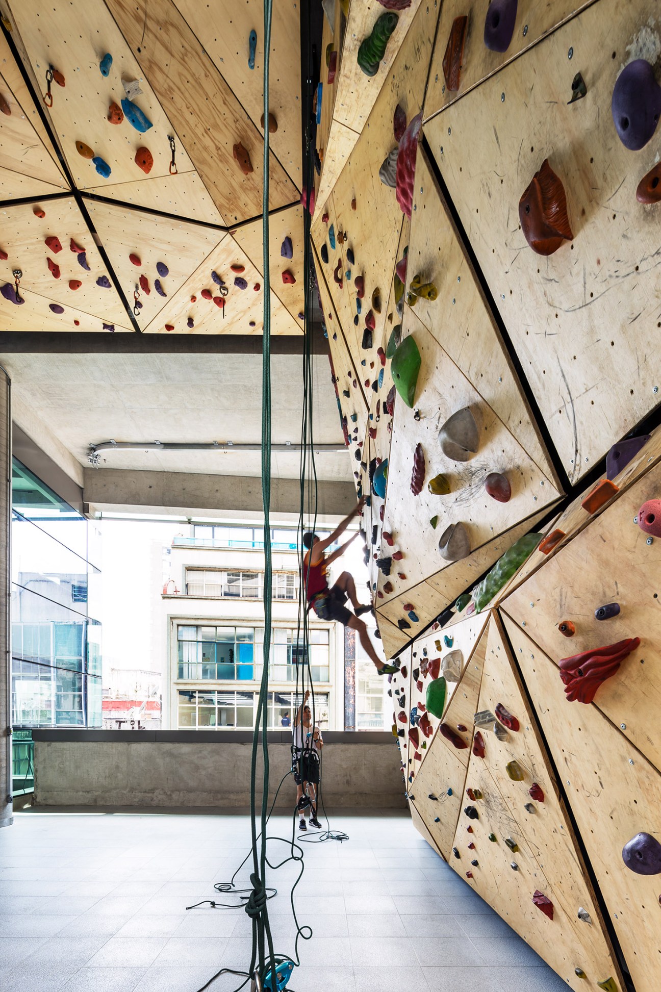 Climber ascends a tall wooden climbing wall with colorful holds at Sesc 24 de Maio in São Paulo.