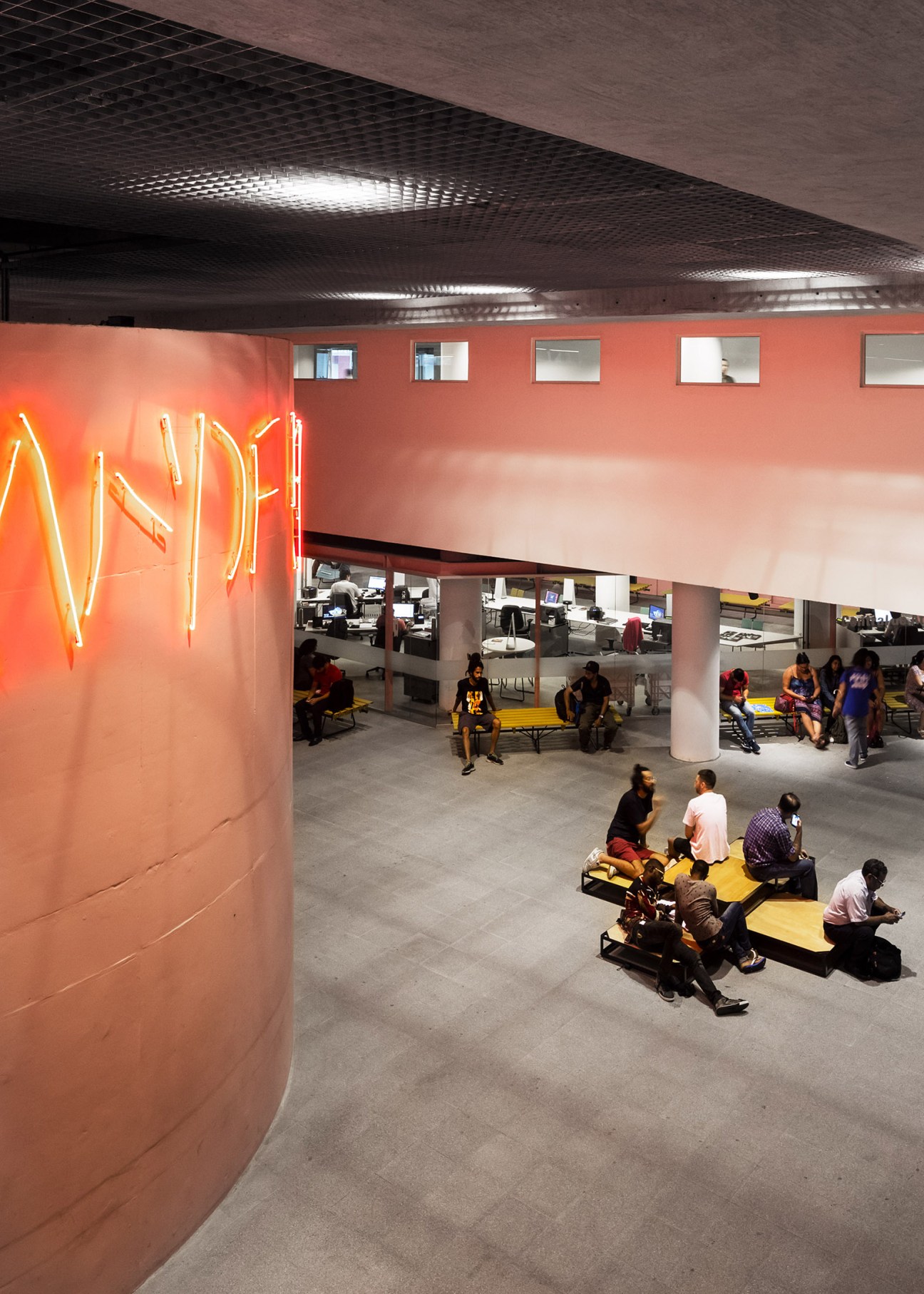 People gathering on yellow benches in a bright open-plan space with coral-red walls and neon signage at Sesc 24 de Maio.