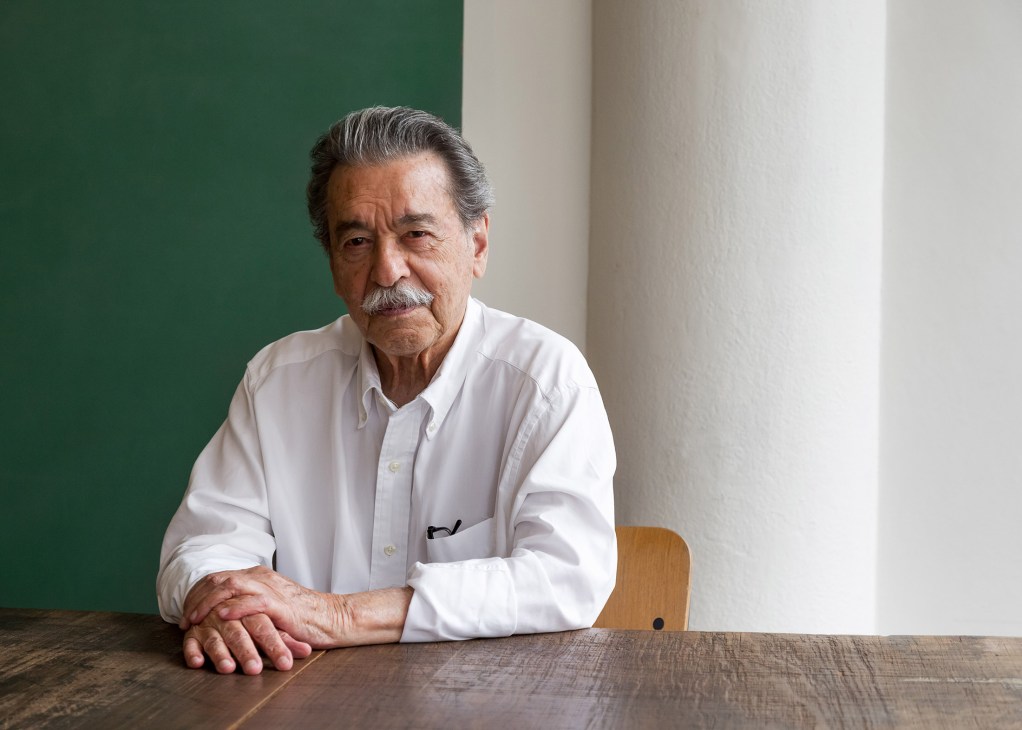 Architect Paulo Mendes da Rocha, aged 89, seated at a wooden table wearing a white shirt against a green wall.