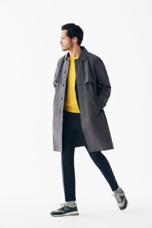 Man wearing grey coat, yellow jumper, dark trousers and New Balance trainers against white background.
