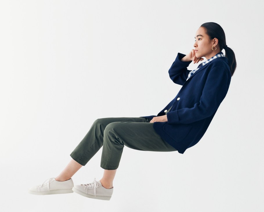 Woman wearing navy blazer, checked shirt, olive trousers and white trainers, seated against white background.