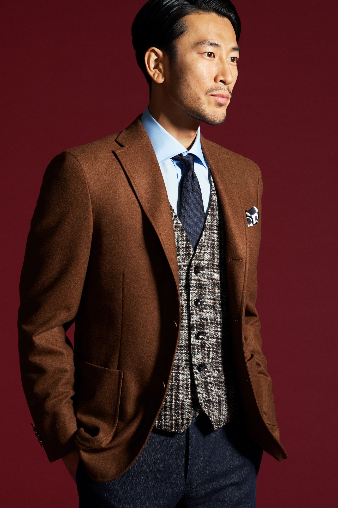 Man in brown Boglioli jacket with grey waistcoat and navy tie against maroon background